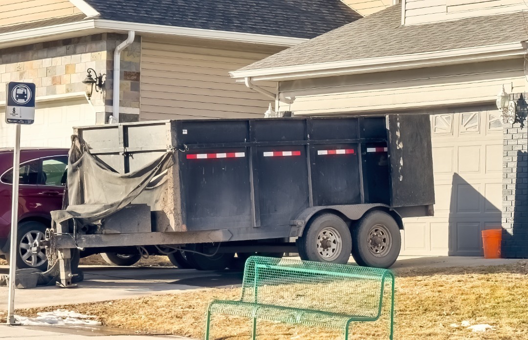 A black dump trailer with a tarp is parked in a residential driveway, likely used for hauling construction debris or yard waste.