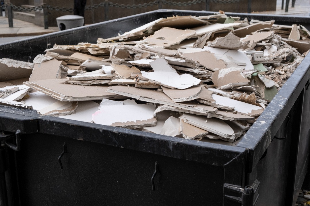 A large dumpster filled with broken drywall and construction waste sits on a city sidewalk after a renovation project.