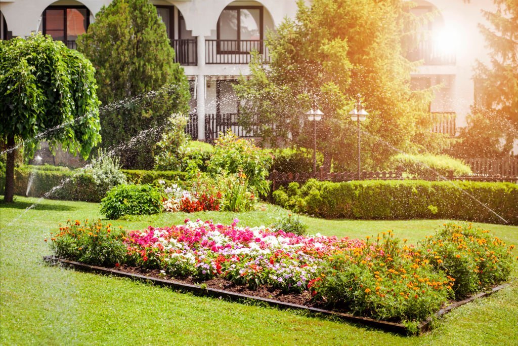 A lush garden with colorful flower beds and green grass is watered by sprinklers in front of a modern residential building.