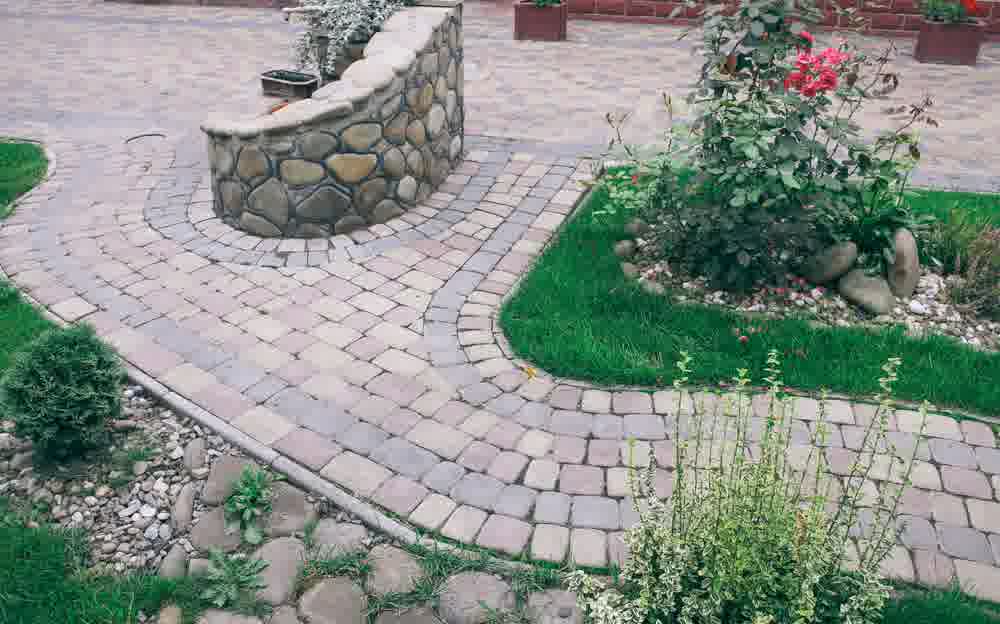 Decorative stone paver walkway curves through landscaped yard with green grass, rose bush, and round garden bed with edging.