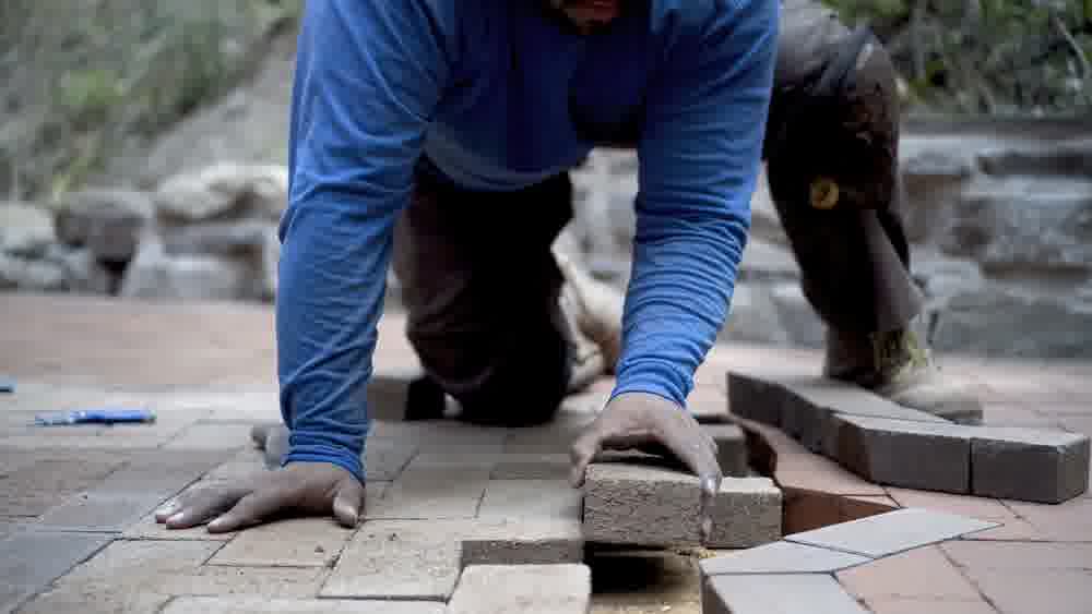 Worker installs brick pavers by hand on outdoor patio, fitting each piece tightly in sand base for a durable and level surface.