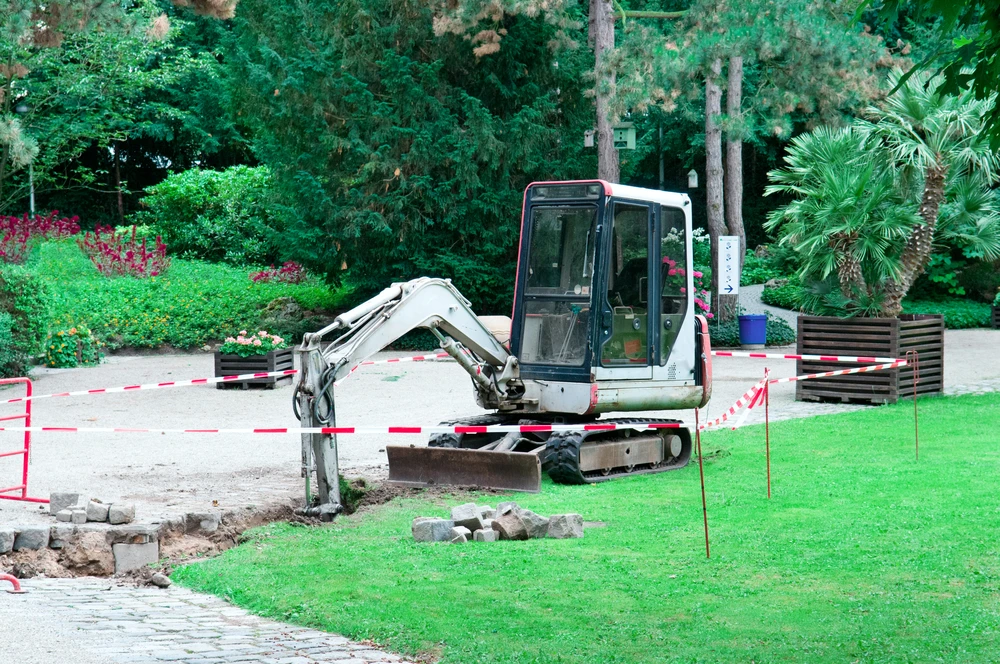 Mini excavator handles concrete demolition along a taped off park walkway.