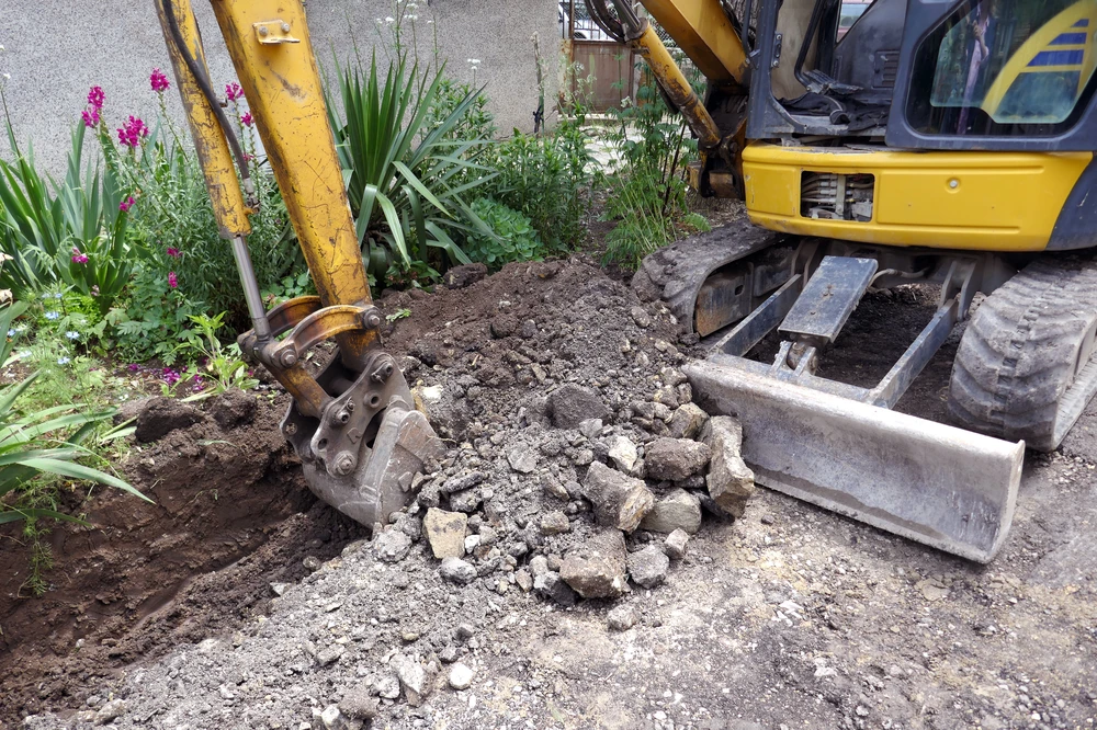 Yellow excavator breaks concrete beside a garden during concrete demolition.
