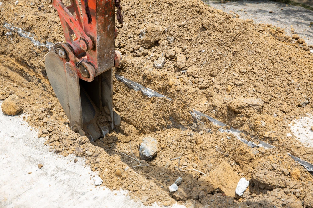 Excavator bucket digs into pavement and dirt during concrete demolition work.