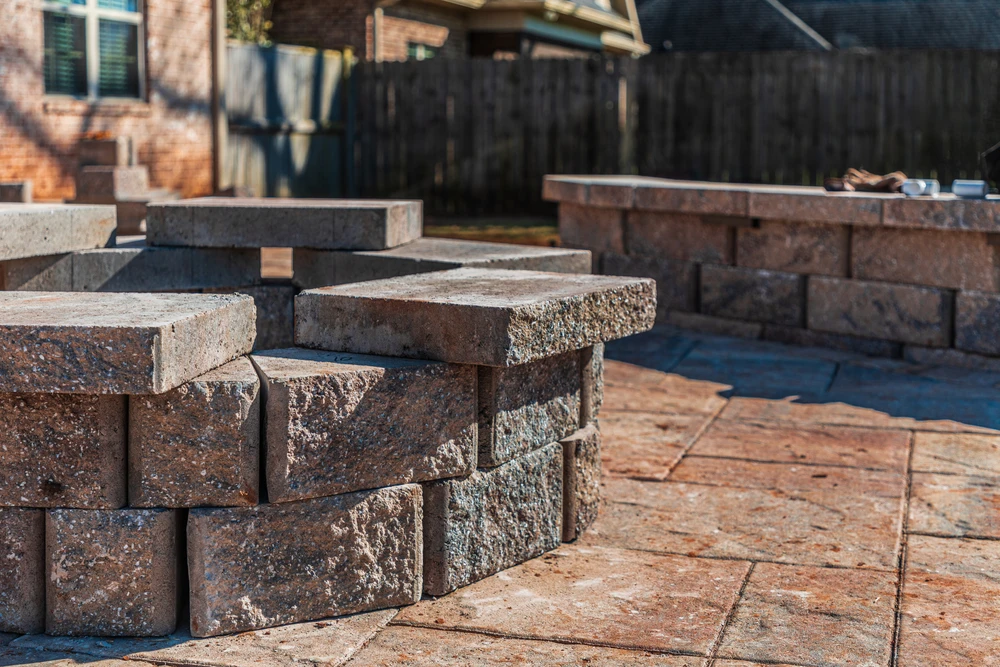 Stacked brown and gray masonry blocks sit on a warm-toned paver patio during landscape construction beside a low stone wall.
