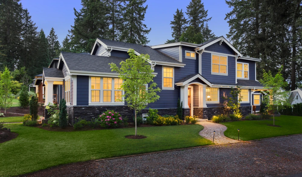 Landscape construction frames a navy blue home with curved stone paths, green lawn, young trees,s flower beds, and warm lighting.