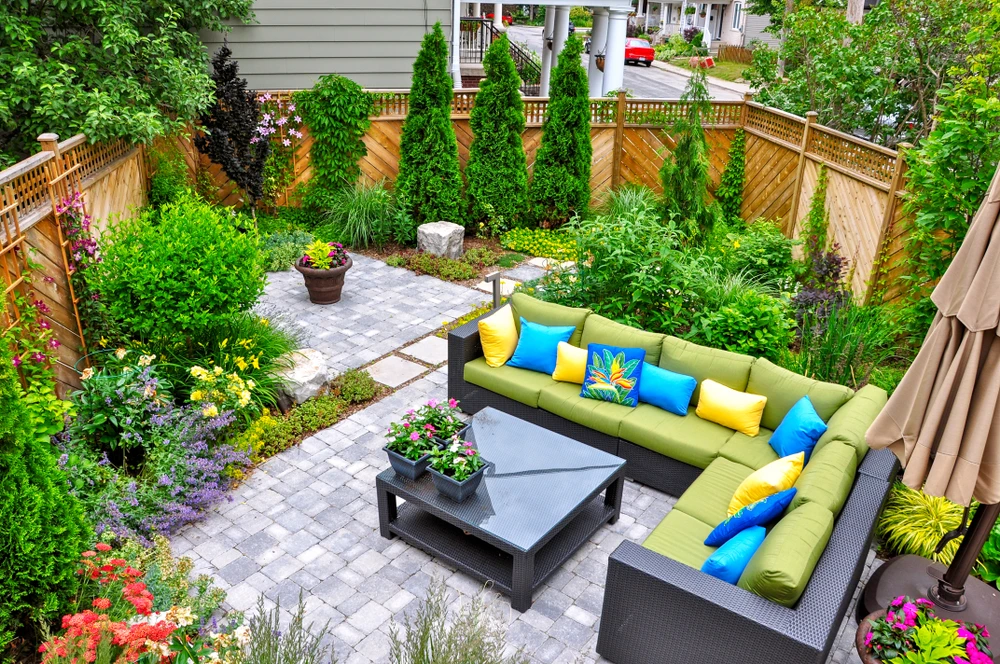 Landscape construction creates a lush backyard patio with gray pavers green seating bright pillows flowers and a wood fence.